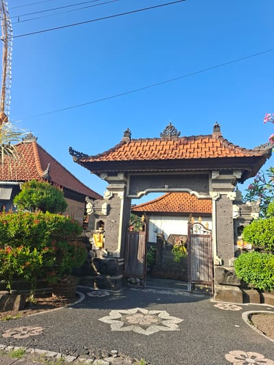 The image shows an ornate stone gateway with a tiled roof, likely part of a traditional Balinese compound, in the village of Blahbatuh, Indonesia. The gateway is constructed from dark, rough-hewn stone blocks, topped with a dark wooden frame and an orange-tiled, multi-tiered roof. Decorative carvings adorn the top of the gateway structure, and two weathered wooden gates, one partially open, stand within the archway.
To the left of the gateway, a lush green tree with red-tinged leaves is planted in a woven basket. Behind the tree, a building with a similar orange-tiled roof is visible, its exterior walls appearing to be a lighter color. The pathway leading up to the gate is paved with dark gravel and features inlaid patterns of light-colored stone creating floral motifs.
To the right of the gateway, another well-maintained green shrubbery is present, partially obscuring the stone structure. In the background, beyond the gateway, another building with an orange-tiled roof is visible. The scene is bathed in bright sunlight, casting clear shadows, and the sky above is a vibrant, cloudless blue. There are no people or animals visible in the image. The overall impression is one of a well-kept, culturally rich entrance to a residential or religious compound.