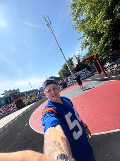 The image is a selfie taken on an outdoor basketball court with a red and black playing surface, located in Vladivostok, Russia. The basketball hoop and backboard are visible in the background, along with a chain-link fence and trees. A man in a blue basketball jersey with the number "55" is in the foreground, smiling at the camera. He has tattoos on his arm. In the background, another person is standing near the basketball hoop. There is graffiti on the wall behind the basketball court, some of which appears to be advertising. A tall cell tower is visible in the sky.