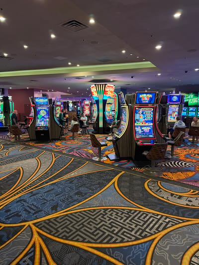 The image depicts the interior of a casino floor in Las Vegas, United States of America. The scene is characterized by numerous brightly lit slot machines arranged in rows and clusters across a vast carpeted area. The machines feature large digital screens displaying various game titles such as "METAL REBELS" and "SAMURAI 888," with prominent progressive jackpot amounts, including "$12,835.42" and "$16,497.90."
The floor is covered with an intricately patterned carpet predominantly featuring shades of blue, gray, and black, accented by sweeping orange and yellow lines, and maze-like geometric designs. Overhead, the ceiling is a light color, equipped with recessed circular spotlights, square ventilation grilles, and some linear ambient lighting casting soft green and purple hues in sections.
Approximately ten to twelve adults are visible within the frame. Most are seated on dark, adjustable stools directly in front of slot machines, actively engaged in play. Specific individuals include: a bald male with a tattoo on his left arm, wearing a dark t-shirt and shorts, standing by a machine on the far left; two males standing between machines in the mid-ground, one in a green long-sleeved shirt and another in a dark t-shirt; a female in a patterned top and a male in a light blue hooded sweatshirt both seated at machines in the mid-ground right; and a female with light hair and a male in a light blue polo shirt seated at machines in the background right. The overall atmosphere is that of a moderately populated casino environment.