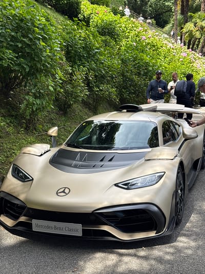 The image shows a tan Mercedes-Benz Classic car parked on a paved surface, likely a road or driveway, adjacent to lush green foliage consisting of bushes and trees. Several people are standing behind the car, their actions obscured but possibly observing or admiring the vehicle. The setting is inferred to be in the city of Como, Italy, based on the prompt’s context. The car's front end is prominently displayed, highlighting its grill, headlights, and the "Mercedes-Benz Classic" license plate. The overall environment suggests an outdoor setting, possibly a garden or park area. The orientation of the image is vertical.