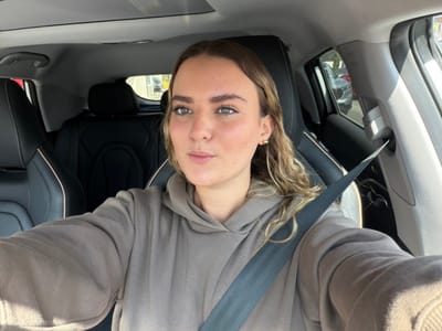 A young woman is captured in a close-up selfie inside a car, looking directly at the camera with a neutral expression. She has light-colored, possibly blonde or light brown, wavy hair that frames her face. She is wearing a light brown or grey hooded sweatshirt and has a gold-colored hoop earring visible in her right ear. A dark seatbelt crosses her chest and shoulder, with the word "AIRBAG" faintly visible on what appears to be part of the seat or pillar near her left shoulder.
The setting is the interior of a car, likely the front passenger seat, during daylight hours, indicated by the bright, clear lighting. The car's upholstery is dark, possibly black leather or fabric. Through the car windows, reflections of other vehicles and an urban or suburban outdoor environment are visible, suggesting the car is either parked or stationary. The image implies a sunny day. Given the location context, this scene is taking place in Puebla City, Mexico.