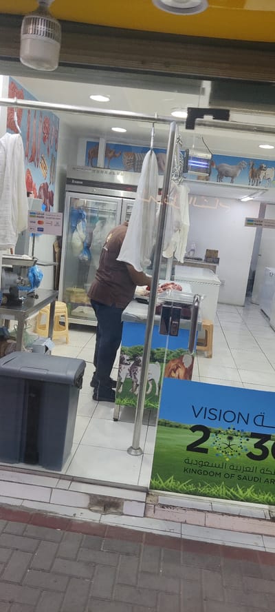 A man in a brown shirt is seen cutting meat behind a glass display in what appears to be a butcher shop in Jeddah, Saudi Arabia. The shop window features posters with animal imagery and the "Vision 2030" logo of the Kingdom of Saudi Arabia. Inside the shop, there is a meat grinder, a meat cooler with hanging carcasses, and various tools and equipment. White garments hang from a metal bar near the front of the shop. The scene is viewed from the exterior, with a grey trash can visible near the entrance and a view of the sidewalk outside.