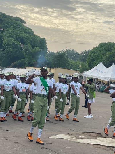 In Alagba, Nigeria, a group of young adults in matching green uniforms and white shirts with "NYSC" printed on them are marching outdoors. The uniforms include green caps, green trousers, white gloves, and orange boots. Some individuals are wearing green sashes over their shirts. One person in the foreground, facing left, is saluting with their right hand, their mouth slightly open as if giving a command. Another person, to the right and slightly behind, is holding up a pink phone, appearing to take a picture or video of the marching group. In the background, white tents are set up, and a truck is visible. Trees and power lines are present in the upper portion of the image, against a cloudy sky. The ground is paved with asphalt.