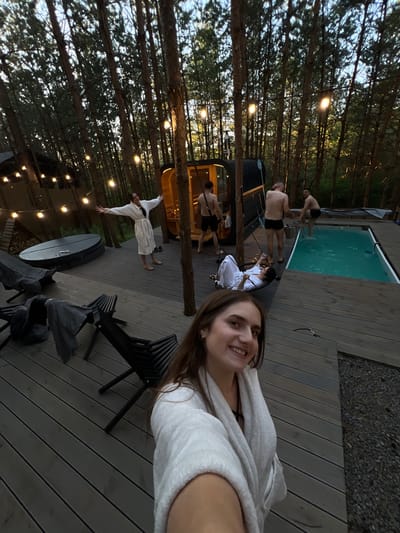 The image depicts a group of people on a wooden deck amidst a pine forest. In the foreground, a young woman with long brown hair, wearing a white robe, smiles at the camera. Behind her, a woman in a white robe is lying on a hammock chair. To the right, three men are visible. One man, wearing black swim trunks, is stepping into a small swimming pool. Another man, also in black swim trunks, stands near the pool. A third man, wearing black shorts, is near a wooden sauna structure. String lights are visible in the background, illuminating the trees. A dark-colored hot tub is situated on the deck to the left. The overall setting suggests a recreational area in a natural, wooded environment, possibly a resort or private retreat. The image was taken in Ivachiv Horishnii, Ukraine.