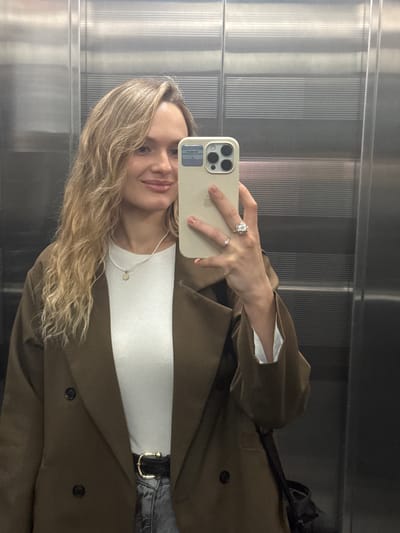 The image shows a young woman taking a mirror selfie in what appears to be an elevator. She is wearing a brown blazer over a white top and jeans. She has blonde hair and is smiling. Her right hand is holding a phone to take the picture, and she is wearing two rings on her left hand. The elevator walls are made of corrugated metal, which reflects the scene. The overall mood is casual and confident. There is no visible text in the image. The location context of Irkutsk, Russia is not visually evident from the image itself.