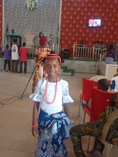 A young girl stands smiling in a room in Yelwa, Nigeria. She is adorned with traditional attire including a white top, a blue and silver patterned wrap skirt, a coral necklace, and coral beads in her hair and on her wrist. In the background, there is a stage with chairs and people standing behind it, including individuals wearing sports jerseys. To the right, people are seated in chairs. A screen is mounted on a patterned wall. The setting appears to be inside a church or event hall.