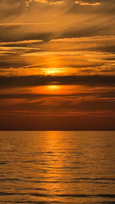 The image depicts a sunset over the Baltic Sea, likely from the coast of Jūrmala, Latvia, given the warm, golden hues and the characteristic appearance of the sea and sky at this time of day. The sun, a bright orb of orange light, is partially obscured by layers of clouds that stretch horizontally across the sky. These clouds are painted in various shades of orange, yellow, and muted gray, with some exhibiting wispy, striated patterns. Below the sky, the sea reflects the dramatic colors of the sunset, creating a shimmering, golden surface. The water is calm, with gentle ripples contributing to the textured appearance of its surface. The horizon line is clearly defined, separating the expansive sky from the vast expanse of water. There are no people, structures, or any other man-made objects visible in the frame, emphasizing the natural beauty of the scene. The overall impression is one of serene, tranquil natural beauty.
