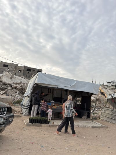 This image depicts an outdoor market scene in Jabalia, Palestinian Territory, set against a backdrop of extensive structural damage.
In the foreground, a dark-colored vehicle, possibly a car, with a damaged front bumper and visible license plate "10 11", is parked on the left. The ground is unpaved, consisting of dry, light brown soil and scattered debris.
Centrally positioned is a makeshift market stall constructed from a white tarpaulin draped over a frame, likely scaffolding or posts. The stall's interior reveals various items on display, resembling produce, with a man inside looking towards the right and gesturing with his right hand. To the left of the stall's entrance, several black containers holding small green plants are arranged on the ground.
In front of the stall, several individuals are present. A person in a dark jacket and light pants stands facing the stall. To their right, two children are visible, one in a striped top and another, younger child in a light-colored, patterned pajama-like outfit, both looking towards the stall's interior. Further to the right, two adults walk away from the stall towards the right side of the frame. One adult wears a light plaid shirt over dark pants and flip-flops, while the other, partially obscured, wears a dark top and lighter pants.
The background reveals a landscape of widespread destruction. Directly behind the market stall and extending to the left, a large pile of concrete rubble and twisted rebar indicates a collapsed structure. Beyond this, partially destroyed multi-story buildings with exposed brickwork and missing sections are visible under an overcast sky with scattered clouds. More rubble and damaged structures are visible on the right side of the frame, behind the market stall, suggesting a wide area has been affected.