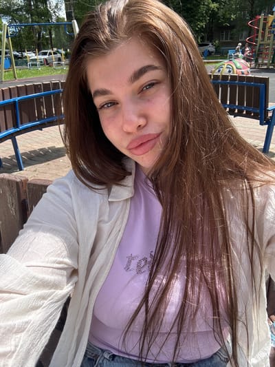 A portrait-style selfie captures a young woman outdoors in what appears to be a park or residential courtyard in Rechytsy, Russia. She has long, straight, dark brown hair, which is highlighted by the bright sunlight, and light-colored eyes. Her expression is neutral with a slight pout.

She is wearing a light purple top adorned with a shiny rhinestone embellishment that forms text or a pattern on the chest, partially obscured by her hair. Over the top, she wears a loose-fitting, light-colored (off-white or cream) collared jacket or cardigan. A denim garment, possibly jeans or a jacket, is visible on her side.

The background reveals a sunny daytime scene with various elements of a playground. Prominent features include blue metal playground structures, a colorful animal-shaped ride-on toy (resembling a ladybug) with red, yellow, green, and blue sections, and a wooden bench with blue supports. Further in the distance, green trees, parts of residential buildings, and a white car are visible. The ground appears to be paved. The image is taken at a tilted angle, common for selfies.