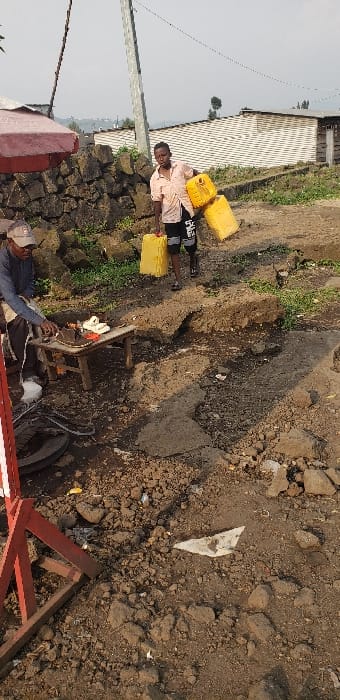 The scene depicts an outdoor environment in Buzove, Democratic Republic of the Congo. The ground consists of uneven, dry earth, loose rocks, and sections of broken concrete or asphalt, with scattered debris.

In the mid-ground left, a male subject, appearing middle-aged, is seated on a low stool. He wears a light-colored baseball cap, a light blue long-sleeved shirt, and dark trousers. His hands are positioned over a low, rectangular wooden table, approximately 0.5 meters in height, where various items, including a dark shoe/sandal and a light-colored sandal, are arranged. Below the table, a dark, circular object resembling a tire rim is visible.

In the mid-ground center, another male subject, appearing younger, walks towards the viewer's left. He wears a light-colored (possibly peach or light orange) short-sleeved collared shirt and dark shorts with white lettering. He is actively carrying three large, rectangular yellow plastic jerrycans: one in each hand and a third tucked under his left arm. The jerrycans appear full.

The background features a rough stone wall constructed from dark, irregular rocks on the left. Behind this wall, green vegetation and distant structures are visible. To the right, a tall, light-colored utility pole stands. Beyond the subjects, the ground rises, revealing simple, light-colored buildings, some with what appears to be corrugated metal roofs. The sky is overcast or hazy. The foreground left includes a red, triangular metal structure.