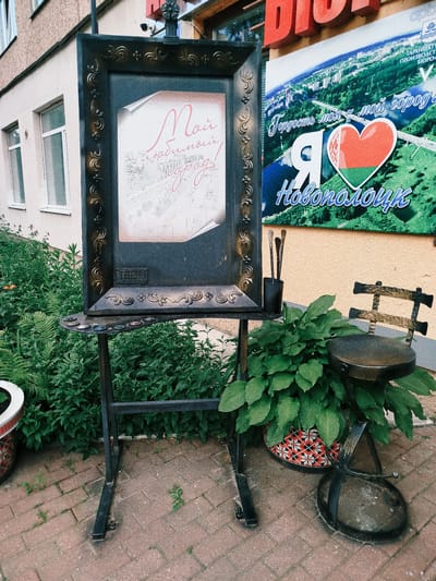 An outdoor easel displays a framed picture with Cyrillic text and a sketch. The text reads "Мой любимый город" (My favorite city). To the right of the easel, a metal chair and a potted plant with large green leaves are visible. Behind the easel and plant is a building facade and a large billboard. The billboard features a green landscape with a river and a city skyline, with text in Cyrillic that includes "Гордость моя - мой город" (My pride - my city) and "Я люблю Новополоцк" (I love Novopolotsk). The scene is set on a brick-paved walkway, indicating an outdoor public space, likely in the city of Navapolatsk, Belarus, given the text on the billboard.