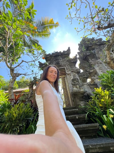 A selfie of a Caucasian woman with long brown hair, wearing a white top, is taken from a low angle in Kedewatan, Indonesia. The background features an ornate, stone Balinese gateway structure overgrown with moss and vegetation. Lush greenery, including trees with broad leaves, and a palm tree with yellow fronds, frame the blue sky. The sun is visible above the gateway, creating a bright, diffused light.