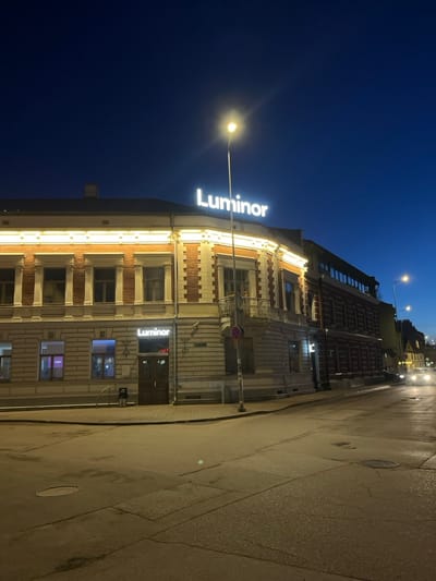 The image displays the intersection of Lielā iela and Jūras iela in Liepāja, Latvia, at night or during deep twilight. The central subject is a two-story corner building located at Lielā iela 11, which functions as a Luminor bank branch.
The building exhibits classical architectural elements, with alternating sections of reddish-brown brick and lighter-toned stucco. Its roofline is illuminated by a continuous strip of warm-toned lights, and a prominent, illuminated white "Luminor" sign is affixed to the roof. A smaller, illuminated "Luminor" sign is positioned above a dark, double-door main entrance on the rounded ground-floor corner. A small, ornate metal balcony protrudes from the second floor of the rounded corner. Windows with light frames are visible across both stories.
To the right, an adjacent, darker brick building extends down Jūras iela. The foreground features a dark asphalt street, appearing damp or reflective, with visible cracks and a circular manhole cover in the lower-left quadrant. Faint street markings are present. A tall street lamp with a bright yellow-white light source stands near the intersection, providing primary illumination. A pole with a circular traffic sign (red circle with a diagonal line) is situated on the sidewalk near the corner building. A dark waste receptacle or bicycle stand is visible near the Luminor entrance. In the distance down Jūras iela, headlights from at least two approaching vehicles are discernible. No individuals are present, and no specific actions or interactions are depicted within the scene.