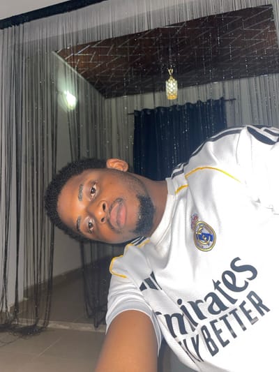 A medium close-up, tilted photograph captures a single male individual of African descent, appearing approximately 20-30 years old, looking directly at the camera with a neutral expression. He has short, dark, curly hair and a short, neatly trimmed dark beard. He is wearing a white short-sleeved Real Madrid jersey with black Adidas stripes on the shoulders and yellow trim. The Real Madrid club crest is visible on the left chest, and the sponsor "Emirates FLY BETTER" is printed in black across the front.
The subject is positioned indoors against a dimly lit background. Above and behind him, a dark ceiling with a reflective, geometric patterned panel is visible, from which a decorative, illuminated gold-toned chandelier hangs. Dark, translucent string or bead curtains are present vertically across the background, partially obscuring what appears to be a darker fabric curtain on the right. A section of a light-colored floor is visible at the bottom left. The image provides no discernible unique geographic markers to confirm the location within Ube, Nigeria; the setting is an unidentified interior space.