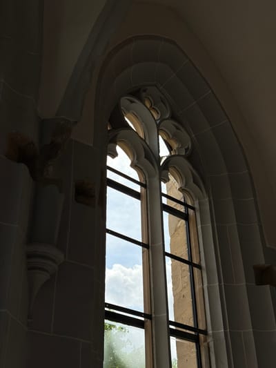 The image shows the interior view of a stone window in Bad Sobernheim, Germany. The window is arched and features decorative gothic-style stone tracery. It is divided by black metal bars, allowing views of the exterior, including a stone wall and glimpses of the sky with clouds, as well as greenery. The stonework around the window exhibits varying degrees of light and shadow, suggesting an interior setting. The window is part of a building constructed from stone blocks. Some of the stonework has signs of wear or missing pieces.