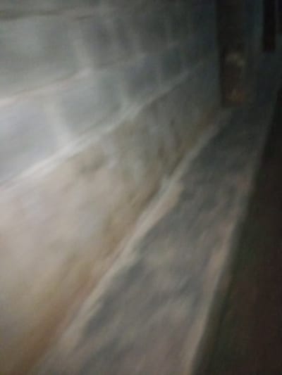 The image depicts an interior view of a basement or crawl space with concrete block walls and a concrete floor. The environment appears damp and unlit, with streaks of discoloration on the walls and floor suggesting moisture or mildew. The perspective is angled, looking down a narrow corridor or channel formed by the walls. In the far right of the image, a blurry object resembling a drum or barrel is visible against the dark wall. The overall scene is dimly lit, with a lack of distinct features, creating a sense of disuse and neglect. The location in Agbara, Nigeria, suggests a possibly industrial or residential underground area.