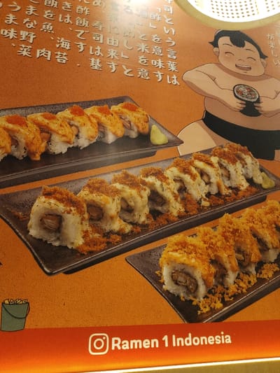 The image shows three different types of sushi on black trays, placed on an orange background. The sushi is displayed on a wall inside a restaurant, presumably Ramen 1 Indonesia, located at coordinates -6.301, 107.278 in Telukjambe, Indonesia. The sushi includes rows of maki rolls topped with various ingredients, including red flakes, white sauce and flakes, and a crispy brown topping. A drawing of a cartoon sumo wrestler is seen on the wall in the upper right of the frame, holding a cup of what appears to be ramen. The image is taken from a low angle, with the trays of sushi tilted slightly upward towards the viewer. Japanese text is visible on the wall in the background.  The photograph likely captures the restaurant's menu offerings.