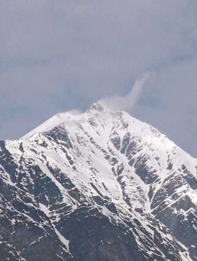 The image depicts the snow-capped peak of a mountain in Dhundi, India, under a cloudy sky. The mountain face is rugged, with visible dark rock formations interspersed with patches of snow. A wisp of cloud or mist is seen blowing across the summit. The lower portion of the mountain appears to have numerous small, colorful objects scattered across its dark surface, which could be prayer flags or debris. The overall impression is a remote, high-altitude environment.