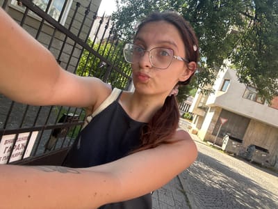 A young woman with auburn hair pulled into a ponytail, wearing large-rimmed glasses and a dark top, takes a selfie in front of a fence and a building on a cobblestone street. She is making a kissing face. In the background, there is a metal fence with a sign in Bulgarian, a building with windows, trees, and two large dumpsters. The sun is casting shadows on the ground. The location is the city of Tryavna, Bulgaria.