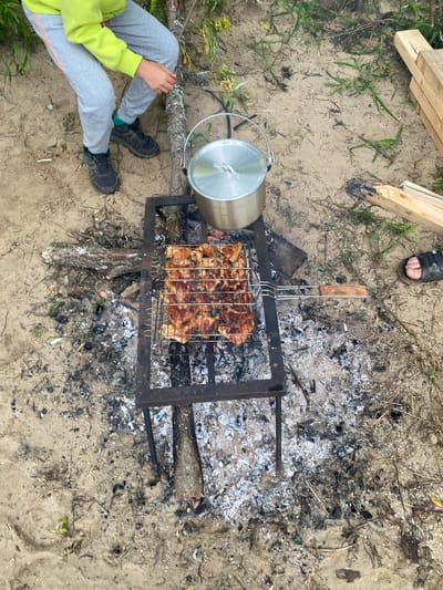 An outdoor scene depicts a person cooking food over a fire. A person, wearing a bright lime green hooded sweatshirt and grey sweatpants with orange stripes, is visible from the waist down, standing in sandy terrain. They are wearing dark athletic shoes and green socks. Their right hand is extended towards a tree trunk.
In the center of the frame, a metal barbecue grill is placed over ashes and burning embers. The grill contains several rows of skewered meat, likely chicken, which is browned and appears to be cooking. A metal pot with a lid sits on top of the grill, to the left of the meat.
The ground is covered in sand, scattered with debris from a fire, including ash, burnt wood, and dry leaves. To the right of the grill, a person's foot wearing a black sandal is visible. In the background, there is some green vegetation and a stack of wooden planks. The scene suggests a campsite or picnic area. The location is identified as Ryabinino, Russia.