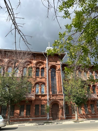 The image shows a brick building with arched windows and decorative elements in Samara, Russia. The building has a pitched roof and appears to be an older, possibly historic, structure. Trees with green leaves are in the foreground, partially obscuring the view of the building. The sky is overcast and dark, suggesting cloudy weather. The street in front of the building is paved, and a silver car is parked on the left side of the image. The overall scene depicts a street view of an old building on a cloudy day in a Russian city.