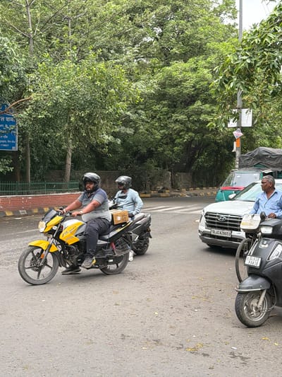 In New Delhi, India, on a street lined with trees and a brick wall, two men ride a yellow motorcycle. The driver, wearing a striped shirt and helmet, steers the bike, while the passenger, in a light blue shirt and helmet, sits behind him holding a cardboard box. In the background, a white car with license plate DL4CAZ8653 is stopped at an intersection. A green truck with a tarp-covered load is behind the car. A black scooter with license plate DL6SB R4438 is also parked nearby, and a man in a blue shirt stands next to it.