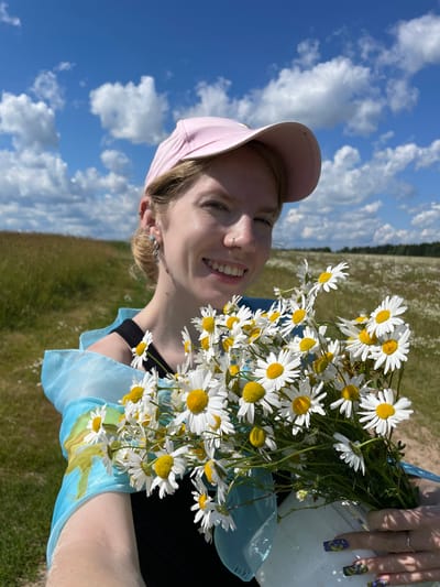 In a sun-drenched outdoor setting, a young Caucasian woman with blonde hair, wearing a pink baseball cap, a black top, and a sheer blue scarf, smiles at the camera while holding a bouquet of daisies. She has multiple ear piercings and a nose ring. The background shows a grassy field dotted with more daisies, and a bright blue sky with scattered white clouds. A dirt path is visible to the right of the woman. The scene appears to be in a rural or natural area, possibly a meadow, and does not provide any specific visual indicators to confirm it is located in the city of Сям-Можга, Russia.
