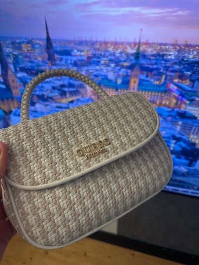 A cream and tan woven Guess handbag with a braided strap is held in a person's left hand in the foreground, obscuring the lower left corner of the image. The handbag features a gold-colored Guess logo and "EST 1981" emblem on its flap. In the background, a blurred image of Hamburg, Germany, at dusk is displayed on a screen. The city skyline is illuminated with vibrant blue and purple lights, highlighting various buildings and structures, including a prominent church spire. The overall scene suggests a product demonstration or personal showcase of the handbag against a digitally rendered backdrop of the city.