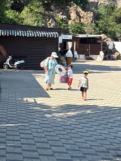 In Partenit, Ukraine, on a sunny day, a woman in a light blue striped dress and a white hat walks along a paved walkway. She carries a pink bag over her shoulder and has an inflatable ring with a colorful pattern in her hands, suggesting a trip to the beach or a swimming pool. To her left, a white scooter is parked next to a dark, corrugated metal building with a striped awning.
Further up the walkway, two children are visible. One child, wearing a yellow hat, white t-shirt, and dark shorts, stands with their legs slightly apart. Another child, in a white t-shirt and red shorts, is walking and holding a colorful inflatable ring. Behind them, a rocky hillside with green foliage slopes down. Structures built into the hillside, possibly shops or shelters, can be seen.
The paving stones on the walkway cast long shadows, indicating it is likely late afternoon. The overall atmosphere appears to be casual and relaxed, consistent with a leisure activity on a warm day. No visible text is clearly discernible in the image, apart from a faded sign on the corrugated building that contains Cyrillic lettering.