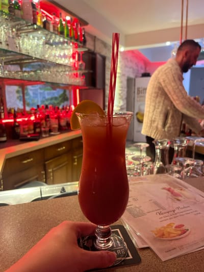 The image captures an indoor bar scene in Meisenheim, Germany, illuminated by prominent red ambient lighting. In the immediate foreground, a human hand with a reddish tint holds a dark circular coaster. Resting on the coaster is a clear hurricane glass filled with a red-orange cocktail containing ice cubes and garnished with a wedge of orange. A red and white striped straw is inserted into the drink. To the right of the cocktail, on a light-colored speckled bar counter, a white menu with German text, featuring "Knusprige Nachos" and an image of fries, is visible. Several inverted, clean drinking glasses are stacked and arranged on the counter. In the left background, a bar area displays multiple liquor bottles on glass shelves, with reflections evident on the surfaces, all bathed in red light. In the right background, a bearded male individual wearing a light-colored cable-knit sweater is partially visible from the chest up, positioned behind the bar. Behind him, a white refrigerator or cooler with the word "Bitter" visible on its side is present. The walls throughout the establishment appear to have a textured, stone-like finish.