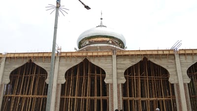 The image shows the under-construction facade of a mosque in Uteunreutoh, Indonesia. The building features a large, reflective dome with a spire at its apex, as well as three large, arched openings in its concrete exterior. These openings are currently filled with scaffolding made of bamboo poles. A utility pole with a spiky antenna extends vertically on the left side of the frame, and a streetlamp is visible in the upper left corner. The sky is overcast. Several people are visible in the lower right corner, some wearing caps. The scene is a construction site, with unfinished concrete, exposed rebar, and wooden formwork visible.