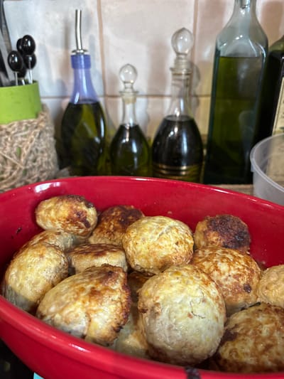 The image shows a close-up view of cooked meatballs in a red bowl, placed to the right of several green glass bottles. The meatballs are golden brown with some darker, slightly charred edges, indicating they have been pan-fried or baked. They are roughly spherical in shape and piled together in the bowl.
To the left of the bowl, several green glass bottles are arranged vertically. One bottle in the foreground has a purple spout with a silver tip, suggesting it might contain oil or vinegar. Below this bottle, a pair of black olive-shaped objects with white stems are visible, possibly part of a utensil set or food decoration. The bottles appear to be on a tiled surface, with a textured material, possibly burlap or twine, visible at the bottom left of the image.
The lighting suggests an indoor setting, possibly a kitchen or dining area. There is no indication of the time of day or weather conditions. The image is a still photograph and does not depict any specific event or activity beyond the presentation of the food and bottles. No visible text is present in the image. The overall impression is one of food preparation or serving.