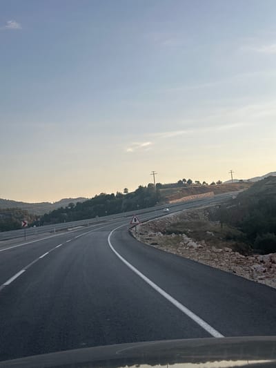 The image shows a road curving through a hilly, rural landscape. The road is paved and has white lane markings. On the left side of the road, there are guardrails and a red and white curved arrow sign. On the right side of the road, there is a triangular warning sign with a symbol of a vehicle going around a curve and a silhouette of a person. The landscape is covered with trees and rocks, and in the distance, there are hills and a clear sky. A white car is visible driving on the road in the middle distance. Power lines and poles are also present in the background. The scene appears to be located in Seydikemer, Turkey, given the context provided.