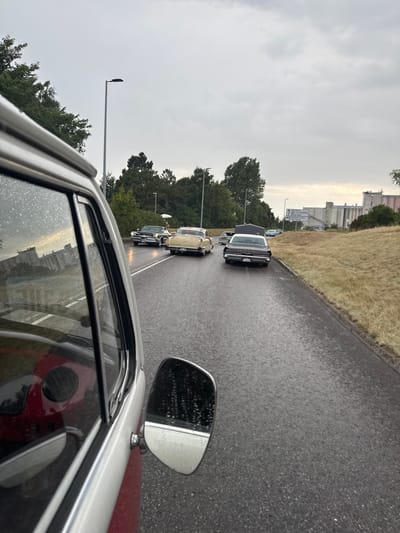 The image captures a convoy of vintage cars driving on a wet asphalt road in Falkenberg, Sweden, under an overcast sky. The perspective is from inside one of the vehicles, looking forward. The foreground is dominated by the interior of the car, including a portion of the dashboard and window, with raindrops visible on the glass. Several classic cars, including a black sedan with a white roof and a cream-colored sedan, are visible ahead on the road. The road is lined with dry, yellowed grass and some trees. In the background, industrial buildings and a signpost indicating "CENTRUM ÅSTRANDEN" and "Falkenbergs Centrum" can be seen. The overall scene suggests a car meet or parade taking place in potentially inclement weather.