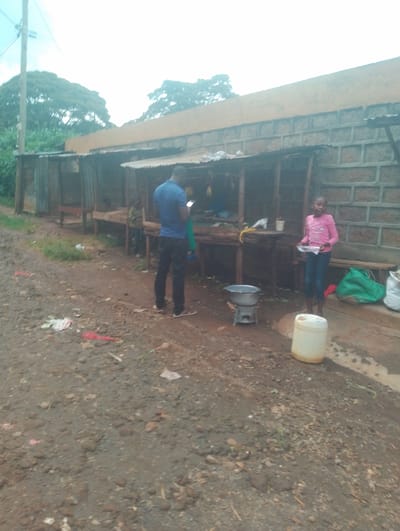 The image depicts a scene outside a small shop in Rungiri, Kenya, located at coordinates -1.244, 36.689. Two individuals, one male and one female, are present. The male, wearing a blue shirt, stands to the left of the frame, appearing to be using a mobile phone. The female, wearing a pink shirt, stands to the right of the frame, holding a white bag. The shop is comprised of a brick structure with a wooden awning.  A few items are visible on the counter, suggesting it may be a small food stall. A large white water container is situated on the dirt ground in front of the stall. The environment is characterized by a dirt road, overgrown grass and several trees in the background.  The image appears to have been captured during the daytime, and the lighting suggests overcast skies.