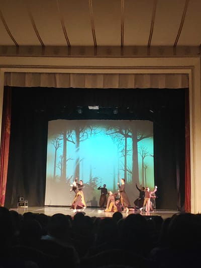 The image shows a stage performance in a theater. Several dancers, dressed in dark, flowing costumes, are performing on a brightly lit stage. The backdrop is a projected image of a forest scene with tall, bare trees and a vibrant turquoise sky. Some of the dancers are in motion, with arms raised and bodies contorted, suggesting a dramatic or expressive dance. Others are positioned on the floor or in more grounded poses. The stage is wide and appears to be made of polished wood. In the foreground, the dark silhouettes of an audience are visible, indicating that the performance is underway. The overall lighting suggests an evening performance. The theater's architecture is visible at the top of the image, with ornate molding and a curtain. The context of Kemerovo, Russia, places this event in a specific geographical location.