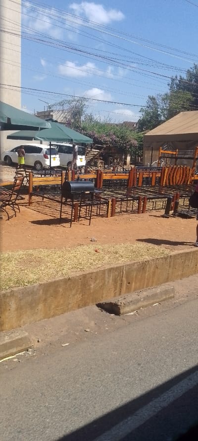 The image captures an outdoor commercial scene in Ngong ward, Kenya, under a clear blue sky with scattered white clouds and numerous overhead power lines. The foreground features a paved asphalt road separated by a concrete curb from a narrow strip of dried grass, which then transitions into a reddish-brown dirt area. This dirt area displays various metal bed frames, some incorporating wooden headboards or footboards, arranged in rows. A black, lidded barbecue grill on a stand is centrally positioned among the bed frames.
To the left, two white vehicles, identified as a hatchback and a van, are parked beneath two large green awnings. A person wearing a light top and a green skirt stands near the left white vehicle. Another person is partially visible near the white van. On the far right, only the lower leg and white shoe of a person are visible, standing near the displayed bed frames. Behind the vehicles and awnings, a light-colored building is visible on the left, and a structure with a brown, tent-like roof is present on the right. The background features green trees, flowering bushes, and additional distant buildings. Shadows indicate direct sunlight.