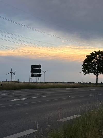 The image depicts a roadside scene at dusk, possibly in Krefeld, Germany, given the context provided. In the foreground, a two-lane asphalt road runs horizontally across the frame, with white lane markings visible. Tall, dry grass lines the right side of the road.
In the middle ground, a row of wind turbines stands silhouetted against the horizon. To their left, a large, dark sign structure with three rectangular panels is prominently displayed. The bottom panel of the sign appears to have graffiti. More wind turbines are visible further in the distance, and a large tree with dense foliage is located on the right side of the frame. The background features a wide, flat landscape under a sky with soft, warm hues of orange and yellow near the horizon, transitioning to cooler blues and grays higher up. Power lines stretch across the sky, supported by poles that are less visible. The overall atmosphere is tranquil and suggestive of an open, possibly rural, area adjacent to a main road.