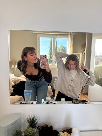 Two young adult females are reflected in a rectangular mirror, positioned inside a residence in Veauchette, France.
The female on the left, with brown hair and bangs, wears a dark brown long-sleeved top, blue jeans with a gold belt buckle, gold hoop earrings, and a gold pendant necklace. She holds a white iPhone with a colorful patterned case in her right hand, angled to capture the mirror reflection. Her eyes are partially closed, and she has a slight smile.
The female on the right, with wavy brown hair parted in the middle, wears a white turtleneck shirt underneath a light beige knit cardigan with a textured pattern and brown buttons, and dark trousers. She is actively styling her hair on the left side of her head with a black electric hair tool held in her right hand, while her left arm is raised. Her expression is focused.
On the white vanity surface in front of the mirror, from left to right, are a white Bose speaker, three small artificial succulent plants in pots, multiple hair scrunchies (including leopard print and solid colors), a small white dispenser bottle, a blue tube of product, a mobile phone lying flat, and a round hairbrush with a patterned handle.
The background reflection visible behind the individuals and through a large, white-framed window shows an interior room with light beige walls. A rumpled bed with light-colored bedding and dark clothing is visible on the left. Outside the window, a clear blue sky with sparse clouds is visible, along with two tall, slender evergreen trees (possibly cypress) and general green foliage, suggesting an outdoor natural setting. A white cabinet or wardrobe is visible on the far right of the reflection.