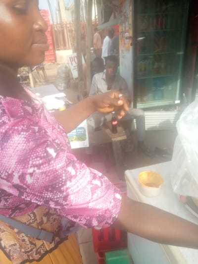 A medium shot captures a woman, partially visible, wearing a pink and white patterned top in the foreground. Her left arm is extended, and she is holding a small white and yellow container. To her right, a glass-fronted refrigerator displays various bottled drinks. Behind the woman, a man is seated on a wooden bench, holding a Coca-Cola bottle. In the background, a busy street scene unfolds with people walking and stalls lined with products. The overall environment suggests a marketplace or a street-side business in Jimeta, Nigeria.