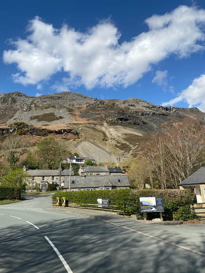 This image captures a view in Llangynog, United Kingdom. A towering slate mountain looms to the left, its rocky, quarried face visible against a backdrop of blue sky with scattered white clouds. A cluster of stone houses with slate roofs lines the roadside. A road runs along the side of the image with white lines and speed ramps are seen along the curb. A sign for the Glendower Holiday Park is affixed to a hedge along the road. The lower-left corner includes foliage, and long shadows are cast across the road.