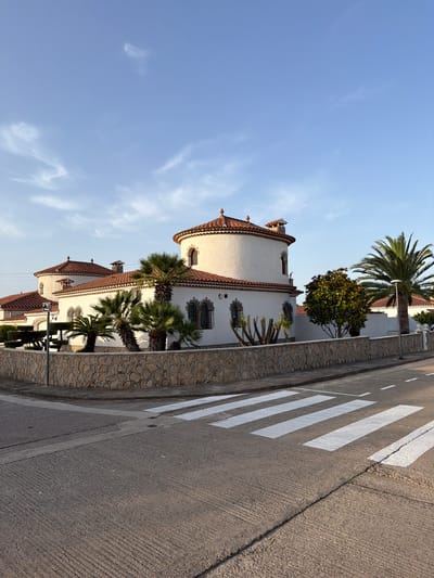 The image shows a street in Mont-roig del Camp, Spain, with a large, white, Mediterranean-style villa on the left side of the frame. The villa has a red-tiled, domed roof and a stone wall surrounding the property. Several palm trees and other greenery are visible within the property walls. A pedestrian crossing is painted on the asphalt road in the foreground, with white stripes. The sky is a clear blue with a few wispy clouds. The scene appears to be in daylight, possibly late afternoon, given the lighting. There are no people or vehicles visible in the image. The area seems to be a residential neighborhood.