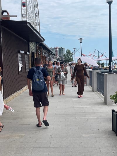 In Gagra, Abkhazia, a man wearing a dark blue t-shirt and dark shorts walks away from the camera on a paved walkway. He carries a blue backpack. Behind him, a line of people walks along the walkway. A woman in a long, dark brown dress and pink sandals walks towards the camera. To the left of the man, a brown building with a dark roof is visible. In the background, a Ferris wheel and modern buildings can be seen. The sky is cloudy.