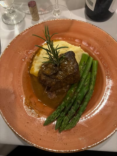 A close-up, overhead shot depicts a culinary dish presented on an orange, speckled plate. The meal consists of mashed potatoes topped with a braised meat item and garnished with a sprig of rosemary. A cluster of green asparagus spears is arranged adjacent to the meat and potatoes. A brown sauce or gravy partially surrounds the dish. In the background, a wine bottle, a cork, and stemware suggest a fine dining experience, but there is no evidence of a specific location within Key Biscayne, United States of America. There are no visible people.