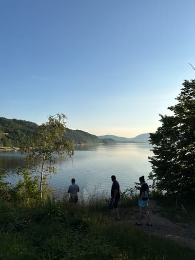 A scenic view of a lake in Lipie, Poland, features three individuals. The lake is calm with gentle ripples reflecting the clear blue sky and distant, tree-covered hills. In the foreground, a narrow dirt path runs along the edge of the lake, bordered by tall grass and vegetation.
On the left side of the path, one person stands facing the water, seemingly absorbed in thought or observation. Further to the right and walking along the path are two other individuals. One, a male, walks ahead, while the second, a female, follows slightly behind, looking at her phone. All three individuals are silhouetted against the bright lake and sky, obscuring specific facial details. The environment is verdant, with lush trees and foliage dominating the landscape. A lone tree with delicate branches stands in the mid-ground, adding to the natural beauty of the scene. The lighting suggests it is either early morning or late afternoon, with the sun casting a soft glow.