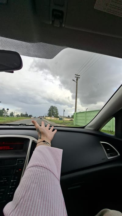 The image is taken from inside a car, looking out through the windshield. A person's arm and hand are visible in the foreground, extended towards the camera, with tattoos on the wrist. The person is wearing a pink and white striped shirt. The car is on a road with a cloudy sky overhead. Power lines and a utility pole are visible in the distance, as is a green fence. The road appears to be a rural or suburban area with some buildings and trees. The location is identified as Smalyavichy, Belarus.