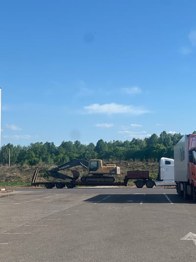 A parked excavator on a trailer behind a semi truck in Kazan, Russia, adjacent to a wooded area and a paved parking lot under a blue sky with scattered clouds.
