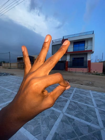 A hand, palm towards the camera, displays a hand gesture in front of a building and sky. Three fingers extend upward, while the thumb and pinky are curled together. The building, two stories, features a white facade, blue windows, and a red accent stripe. A cloudy sky is visible behind the building. A tiled surface with a geometric pattern fills the right foreground.