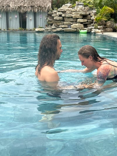 In the city of Nam Hoa Lư Ward, Vietnam, two individuals are submerged in a bright blue swimming pool. The person on the left, a male with long, dark, wavy hair, faces towards the right, his upper body visible above the water. The person on the right, a female with lighter brown hair, faces towards the left, her upper body also above the water. She has a colorful, intricate tattoo on her right arm. Both individuals are interacting with each other, with their hands in the water between them. The pool's surface is rippled, reflecting the surrounding environment. In the background, a rock formation with greenery and palm trees is visible, along with a thatched-roof structure and a bright green inflatable toy in the water. The overall scene suggests a relaxed and recreational setting within the city.