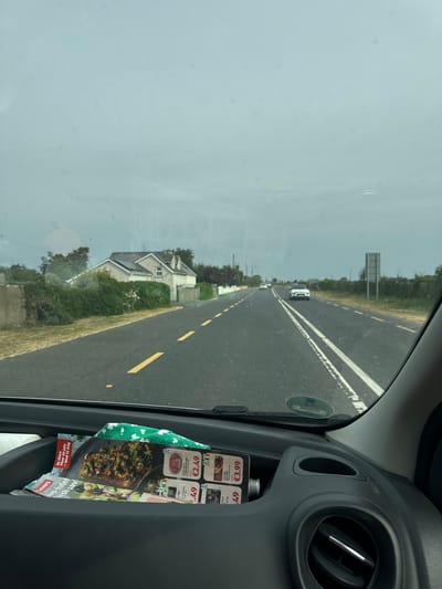 The image is taken from inside a vehicle, looking out through the windshield at a rural road in Killcommon, Ireland. The sky is overcast and a light grey. The road is paved and has yellow dashed lines marking the lanes. There are several white cars visible on the road, some in the distance and one approaching. On the right side of the road, there is a sign and dense green vegetation. On the left side of the road, there is a house with a green hedge in front of it. In the foreground, inside the vehicle, there is a storage compartment on the dashboard containing some leaflets and a green cloth item.